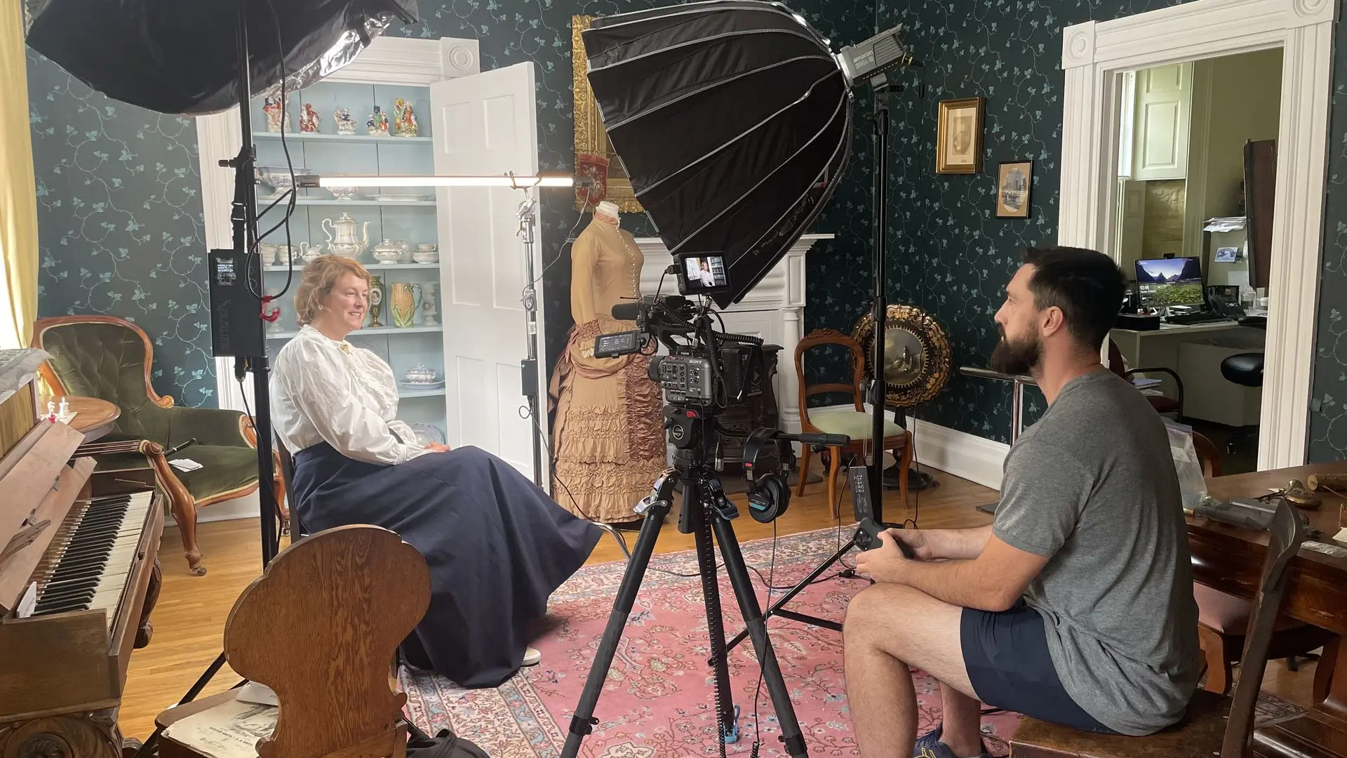 a woman in period clothing is illuminated by studio lights while a director in a grey t-shirt views the scene from behind a camera