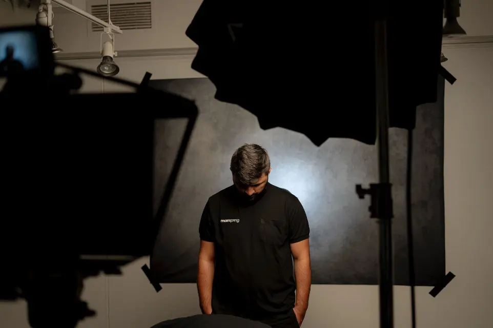 a man wearing a mainspring agency t shirt stands in front of a camera and a light on set