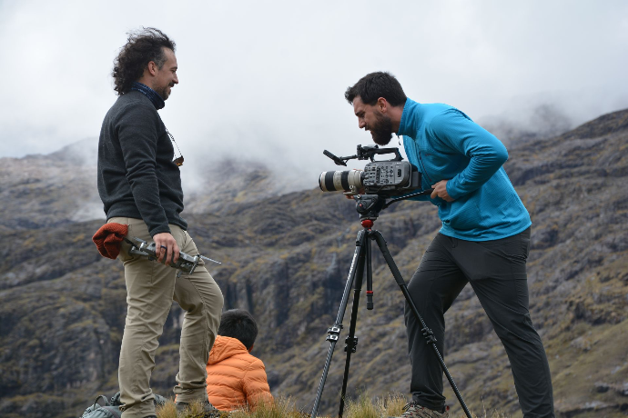 a mainspring agency video crew are filming on top of a mountain, the camera is on a tripod and is being operated by a cinematographer and the production assistant is holding a drone