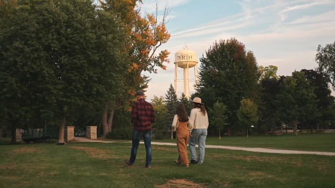 a young, fashionable family walk through Stewart park in Perth, ontario, the water tower in the distance reads PERTH